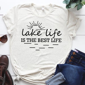 Peut inclure: Un t-shirt blanc avec un motif noir qui dit "lake life is the best life" avec un graphique de soleil au-dessus du texte. Le t-shirt est pos&eacute; sur une surface en bois blanc avec un jean, des lunettes de soleil et des bottes.