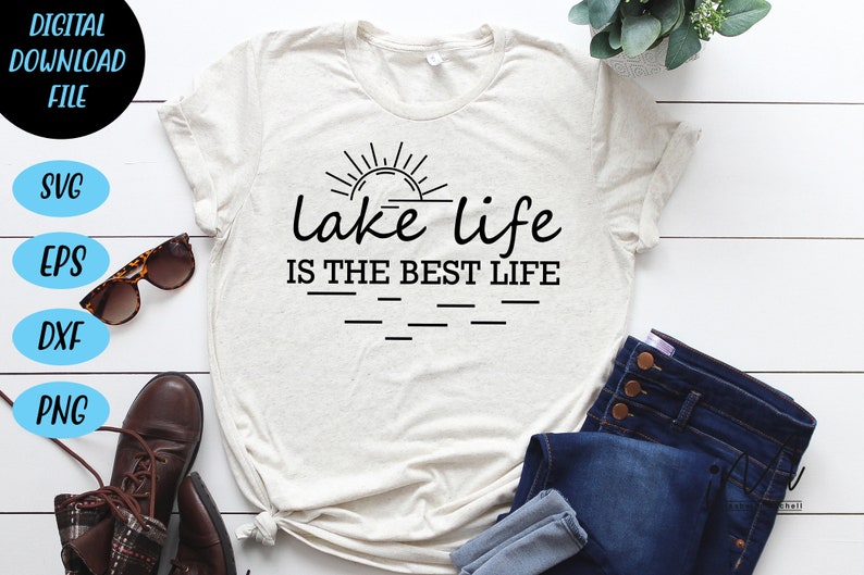 Peut inclure: Un t-shirt blanc avec un motif noir qui dit "lake life is the best life" avec un graphique de soleil au-dessus du texte. Le t-shirt est pos&eacute; sur une surface en bois blanc avec un jean, des lunettes de soleil et des bottes.