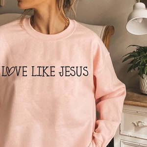 May include: A pink sweatshirt with the text "LOVE LIKE JESUS" in black lettering with a small heart symbol before the word "LOVE".