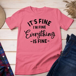 May include: A pink short-sleeved t-shirt featuring black text that reads IT'S FINE I'M FINE Everything -IS FINE- centered on the front. The shirt is displayed alongside a brown hat, a plaid flannel shirt, and a pair of dark blue denim jeans.