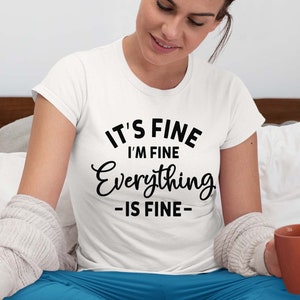 May include: A white short-sleeved t-shirt featuring black text that reads IT'S FINE I'M FINE Everything -IS FINE- in a mix of bold sans-serif and cursive fonts. The shirt is worn with blue pants and a light gray textured cardigan sweater.