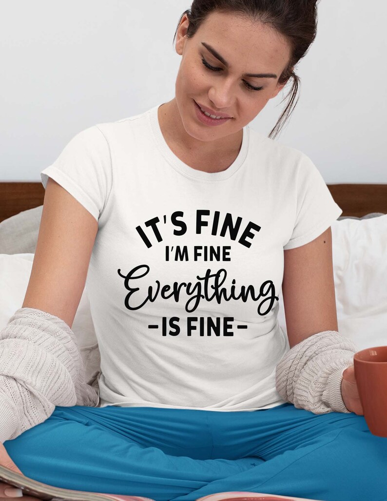 May include: A white short-sleeved t-shirt featuring black text that reads IT'S FINE I'M FINE Everything -IS FINE- in a mix of bold sans-serif and cursive fonts. The shirt is worn with blue pants and a light gray textured cardigan sweater.