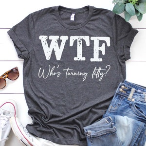 May include: A dark gray t-shirt with the text "WTF Who's turning fifty?" printed in white. The shirt is laid flat on a white wooden surface with a pair of sunglasses, a pair of white sneakers, and a pair of blue jeans.