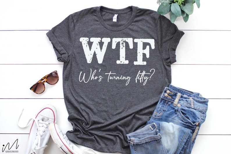 May include: A dark gray t-shirt with the text "WTF Who's turning fifty?" printed in white. The shirt is laid flat on a white wooden surface with a pair of sunglasses, a pair of white sneakers, and a pair of blue jeans.