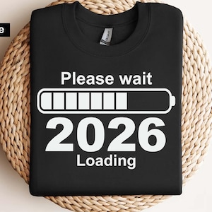 Puede incluir: Sudadera negra con el texto "Please wait 2026 Loading" en blanco. Un gráfico de carga de batería blanco está encima del año. La sudadera se muestra sobre una superficie circular tejida.