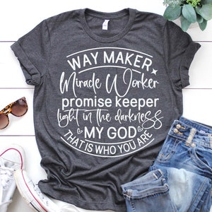 May include: A dark gray t-shirt with a white circle design that says "Way Maker Miracle Worker Promise Keeper Light in the Darkness That is Who You Are My God".