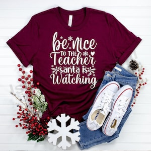 以下が含まれることがあります： 「be nice to the Teacher santa is Watching」の白文字がプリントされた、マルーン色のTシャツ。青いジーンズと白いスニーカーと組み合わせています。装飾要素には、雪の結晶と赤いベリーが含まれています。