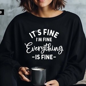 May include: A black long-sleeve sweatshirt featuring white text that reads IT'S FINE I'M FINE Everything -IS FINE- in a mix of block and script fonts. The garment has a crew neckline and long sleeves, with a digital file label in the corner.