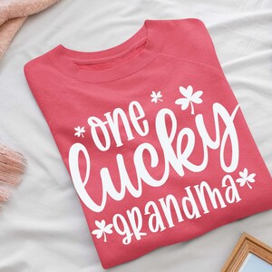 K&ouml;nnte beinhalten: Ein rosa Sweatshirt mit dem Text "one lucky grandma" in wei&szlig;en Buchstaben mit Kleeblatt-Designs.