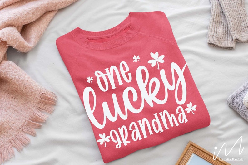 K&ouml;nnte beinhalten: Ein rosa Sweatshirt mit dem Text "one lucky grandma" in wei&szlig;en Buchstaben mit Kleeblatt-Designs.
