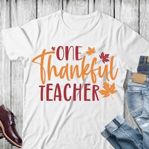 May include: White t-shirt with the text "One Thankful Teacher" in orange and red lettering.  The text is surrounded by small orange and red maple leaves.