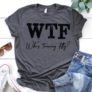 May include: A dark gray t-shirt with the text "WTF Who's turning fifty?" printed in black. The shirt is laid flat on a white wooden surface with a pair of sunglasses, a pair of white sneakers, and a pair of blue jeans.