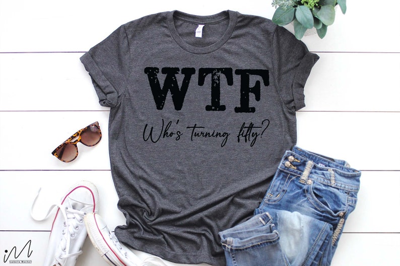 May include: A dark gray t-shirt with the text "WTF Who's turning fifty?" printed in black. The shirt is laid flat on a white wooden surface with a pair of sunglasses, a pair of white sneakers, and a pair of blue jeans.