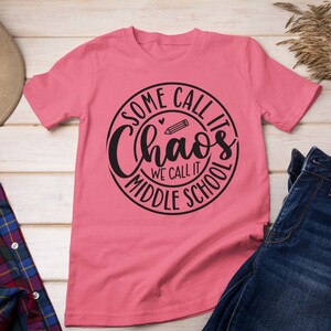 Puede incluir: Camiseta rosa con un dise&ntilde;o circular negro que dice "Some call it chaos we call it middle school" con un l&aacute;piz y un coraz&oacute;n dentro del c&iacute;rculo.