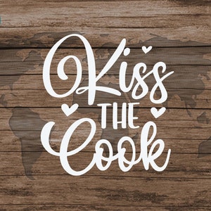 May include: White text on a wood background that reads "Kiss the Cook" with a small heart on either side of "the". The text is in a cursive font. The background is a wood grain pattern with a faint outline of a world map.