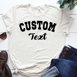 May include: A white t-shirt with a black graphic that says "CUSTOM Text" in a collegiate font.