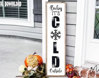 Può includere: Un cartello rettangolare in legno con la scritta "Baby It's Cold Outside" in lettere nere. Il cartello presenta un disegno a fiocco di neve ed è incorniciato in legno marrone chiaro. In primo piano ci sono zucche e zucchette.