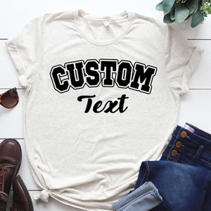 May include: A light beige t-shirt with black text that reads "CUSTOM Text" in a varsity style font.