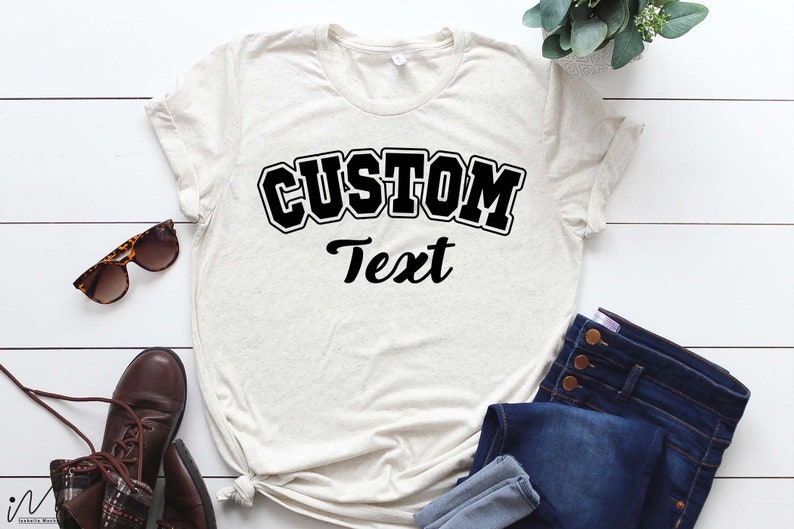 May include: A light beige t-shirt with black text that reads "CUSTOM Text" in a varsity style font.