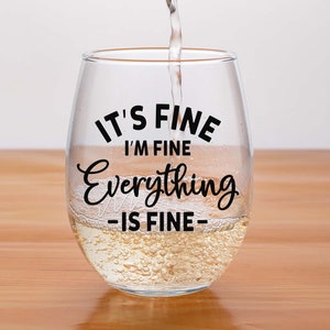 May include: A clear stemless wine glass filled with a pale liquid, featuring the black printed text IT'S FINE I'M FINE Everything IS FINE. The glass has a rounded bowl shape and is being filled with liquid from above, creating small bubbles.