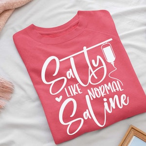Puede incluir: Una sudadera rosa con el texto "Salty Like Normal Saline" en letras blancas. Una bolsa de suero intravenoso blanca se ilustra junto al texto.