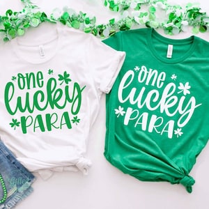 May include: Two white t-shirts with green shamrock designs. The text on the shirts reads "one lucky *PARA*" in green lettering. The shirts are laid out on a white surface with green garland.
