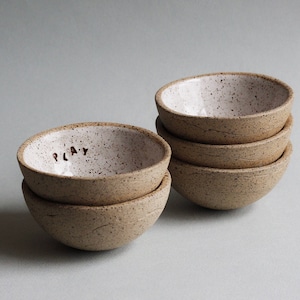 Könnte beinhalten: Ein Set aus fünf kleinen, gesprenkelten Keramikschalen. Die Schale vorne hat das Wort "PLAY" auf der Innenseite.