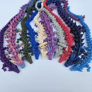 May include: A collection of 10 3D printed dragon figurines in various colors, including purple, pink, green, blue, cream, and red. The dragons are all facing the same direction with their heads raised.