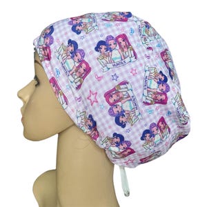 May include: A white and pink patterned scrub cap featuring a repeating design of cartoon characters and the text "HUNTR/X". The cap has a gingham background with musical notes and stars.