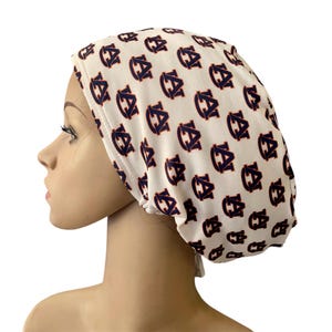 May include: A white scrub cap featuring a repeating pattern of the navy blue and orange Auburn University AU logo. The fabric head covering has a rounded, gathered shape designed to fit securely around the head with a smooth, fitted front band.