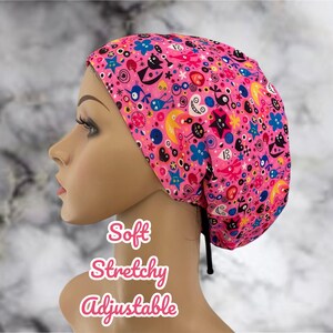 May include: A pink, stretchy, adjustable surgical cap with a colorful, whimsical pattern of eyes, stars, and other abstract shapes. The text "Soft Stretchy Adjustable" is printed below the cap.