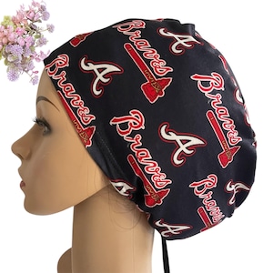 May include: A navy blue scrub cap featuring the Atlanta Braves logo in red and white. The cap has a repeating pattern of the team's name, tomahawk, and the letter "A". The cap is tied at the back.