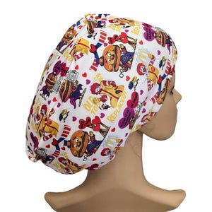 May include: A white scrub cap featuring a colorful repeating pattern of cartoon characters, hamburgers, and red hearts. The fabric includes text elements such as "OLD TOYS CAN BE REPLACED" and "PICTURE US AS VALENTINES... SAY CHEESE" printed in yellow and purple.