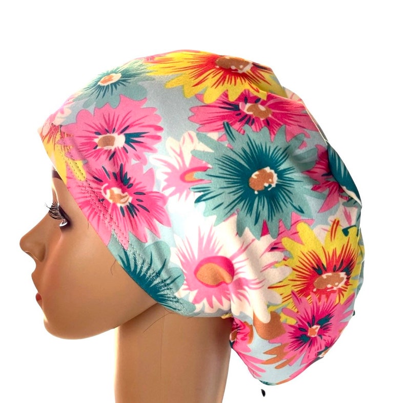 May include: A floral print bouffant scrub cap featuring a pattern of large daisies in pink, yellow, teal, and white. The cap is designed to cover the hair and is made of a soft, lightweight fabric.