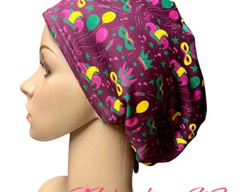 May include: A purple scrub hat with a Mardi Gras themed print featuring green, yellow, and pink colors. The print includes masks, crowns, stars, and balloons.