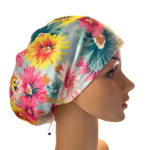 May include: A floral patterned scrub cap featuring large, colorful flowers in shades of pink, yellow, and teal on a light blue background. The cap has a drawstring for an adjustable fit. The design is a cheerful and vibrant accessory.