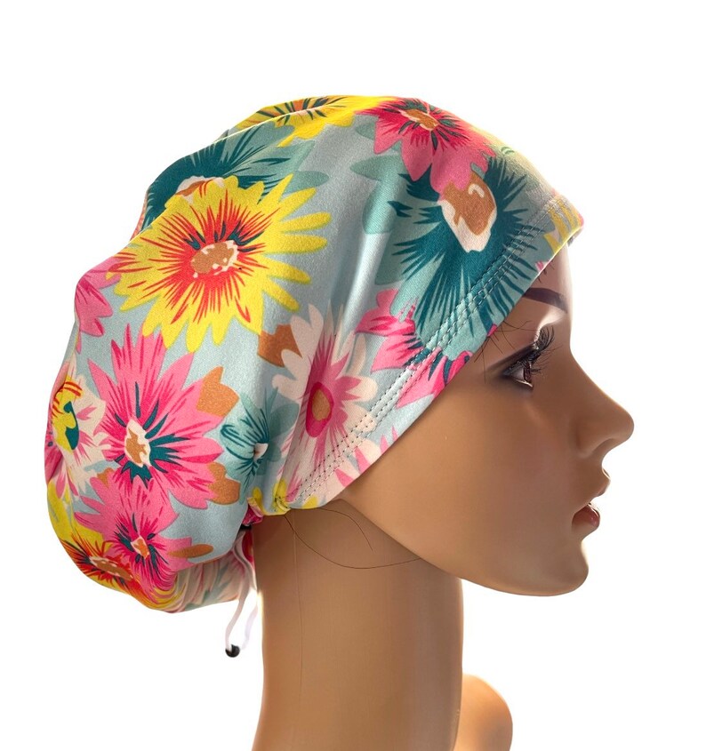 May include: A floral patterned scrub cap featuring large, colorful flowers in shades of pink, yellow, and teal on a light blue background. The cap has a drawstring for an adjustable fit. The design is a cheerful and vibrant accessory.