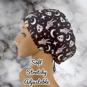 May include: Black adjustable scrub cap with a repeating pattern of white skeleton hands, planchette, crescent moons, and stars. The cap is soft and stretchy. Text on the image reads "Soft, Stretchy, Adjustable."