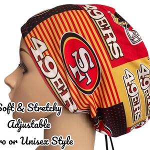 May include: A scrub cap featuring a red, gold, and black pattern with the San Francisco 49ers logo and text. The fabric displays repeating team branding, including helmets and the 49ers name. The cap includes black ties at the base for adjustment.