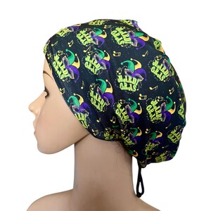 May include: Black Mardi Gras scrub cap with a repeating pattern of jesters, musical notes, and the words "Mardi Gras" in green, purple, and yellow. The cap has a drawstring for an adjustable fit.