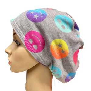 May include: Gray beanie with a colorful smiley face pattern. The beanie has a tie at the back.