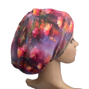 May include: A slouchy beanie hat featuring a vibrant, painterly floral print in shades of magenta, deep purple, orange, and yellow. The fabric displays a soft, smooth texture with a repeating pattern of abstract blossoms set against a dark, moody background.