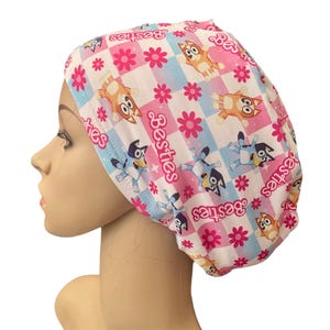 May include: A pink and white patterned scrub cap featuring cartoon characters and the word "Besties". The cap has a checkered design with blue and pink squares, and pink flowers. The cartoon characters are in orange and blue.