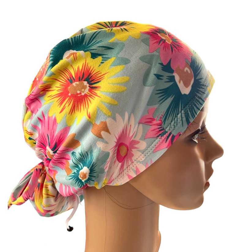 May include: A floral patterned scrub cap featuring a light blue background with large, colorful flowers in shades of pink, yellow, and white. The cap is tied at the back with a drawstring for an adjustable fit. The cap is designed to be worn in a medical setting.