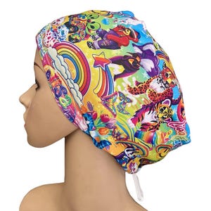 May include: A scrub cap featuring a vibrant, multi-colored print on a yellow background. The pattern includes rainbows, butterflies, stylized animals, and cartoon figures. The cap has a gathered back with a white adjustable drawstring closure.