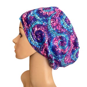 May include: A colorful, patterned cap in shades of blue, purple, and pink. The fabric features a mosaic-like design. The cap is worn on a mannequin head, showcasing its shape and fit. The cap is designed to be worn on the head.