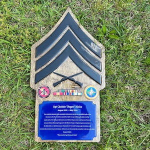 May include: Wooden plaque featuring a black and gray military rank insignia, crossed rifles, and military emblems. A blue plaque with text reads "Sgt Chelsie 'Okurrr' Hicks August 2021 - May 2024". The plaque is set on a grassy surface.