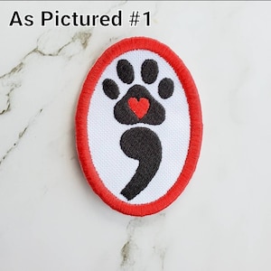 May include: Red and black embroidered patch with a paw print and a semicolon, with a red heart in the center of the paw print. The patch is circular and has a white background.