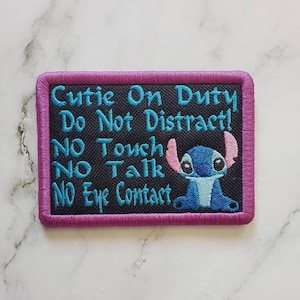 May include: Embroidered patch with a black background and a purple border. The patch features the text "Cutie On Duty, Do Not Distract! NO Touch, NO Talk, NO Eye Contact" in turquoise. A blue cartoon character with pink ears is on the right.