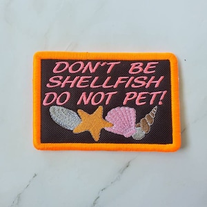 May include: An orange-bordered patch with a black background. The patch has pink text that reads "Don't be shellfish, do not pet!" and features an embroidered image of a starfish and seashells.